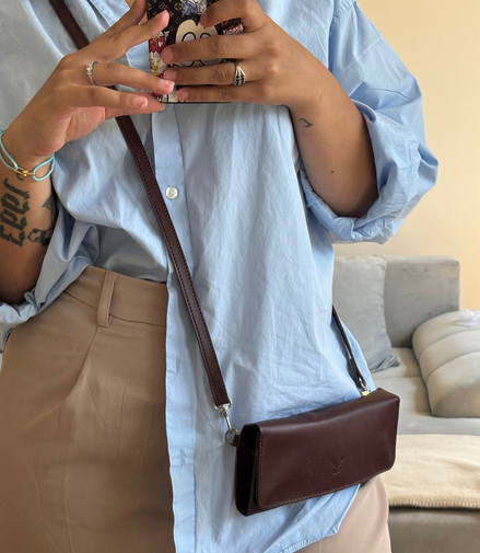 Person wearing a light blue shirt and beige pants, holding a phone with a floral case and carrying markore gul sahar crossbody bag handmade from full-grain leather