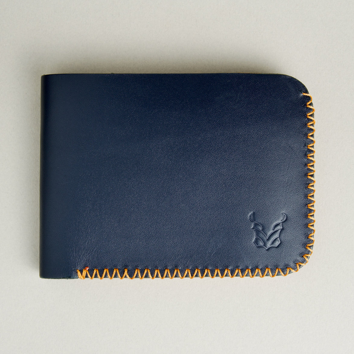 Front view of dark blue full-grain leather bifold wallet featuring handmade amber cross-stitched detailing on the edges and embossed logo. Markore Hunza Edge.