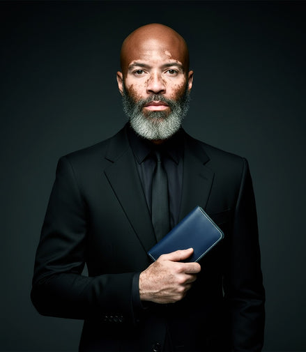 Studio portrait of a distinguished man with a grey beard and Vitiligo on his face wearing a black suit, holding the Markore Karakoram II in Indigo Blue close to his chest. The dark background and sharp lighting highlight both the man’s presence and the refined craftsmanship of the wallet.
