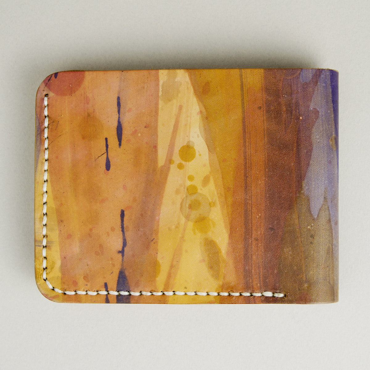 Backside view of hand-painted full-grain leather bifold wallet in purple, amber, and brown, featuring handmade stitching. Markore Canvas Chaos.