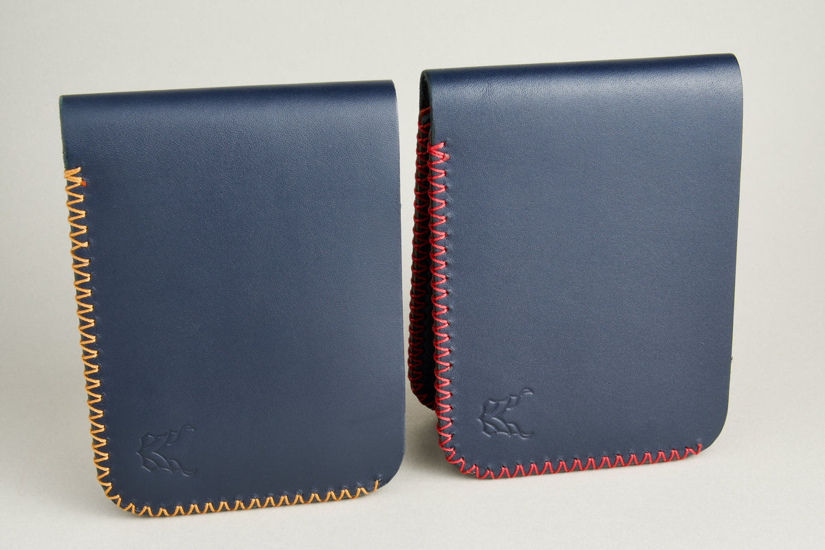 Side-by-side view of two dark blue full grain leather bifold wallets, featuring cross-stitched detailing in amber and red on the edges, and embossed logo. Markore Hunza Edge.