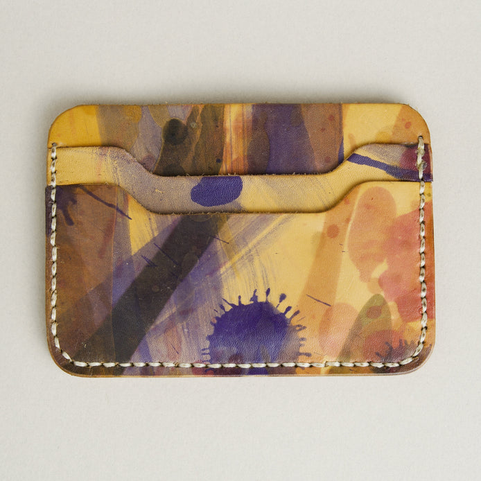 Front side of hand-painted full-grain leather card sleeve in purple, amber, and brown, featuring handmade stitching. Markore Niva Chaos.