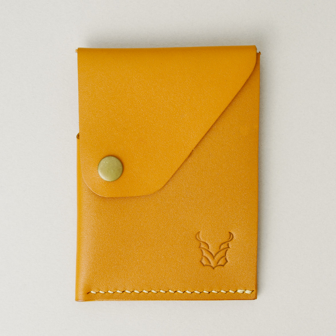 Front view of full grain leather card holder in amber with ivory handmade stitching, featuring a flap with brass press button and embossed logo. Markore Niva Heritage.