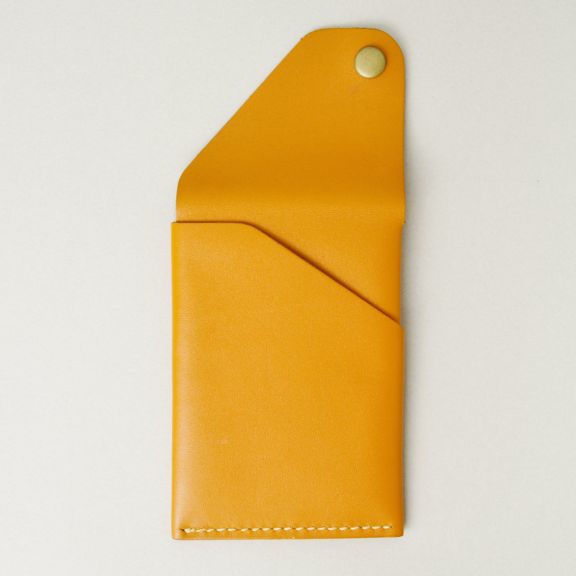 Back view of full grain leather card in amber with ivory handmade stitching, featuring an open flap with brass press button a card pocket. Markore Niva Heritage.