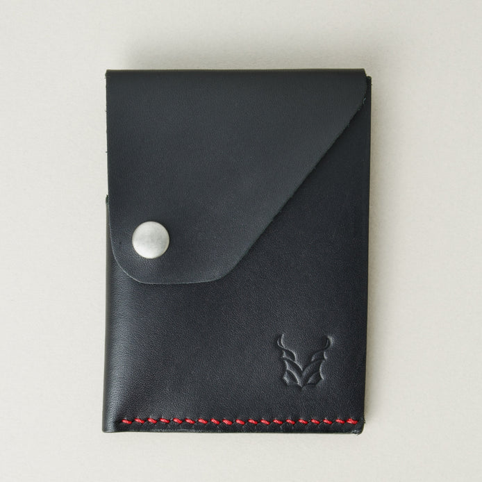 Front view of full grain leather card holder in black with red handmade stitching, featuring a flap with stainless steel press button and embossed logo. Markore Niva Heritage.