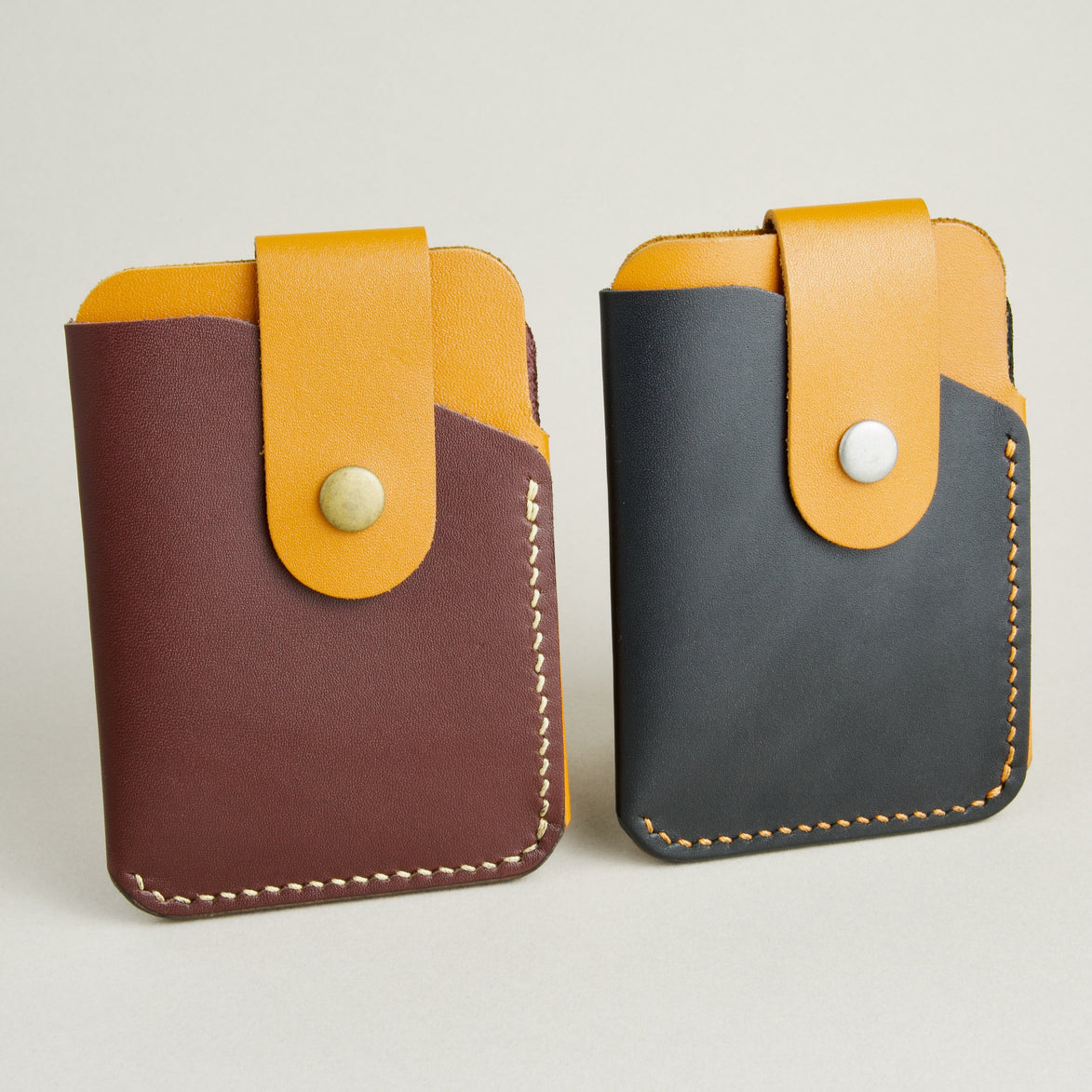 Side view of two full grain leather card holders in midnight black and espresso brown with amber, featuring handmade stitching an a leather loop with press button. Markore Niva Loop.
