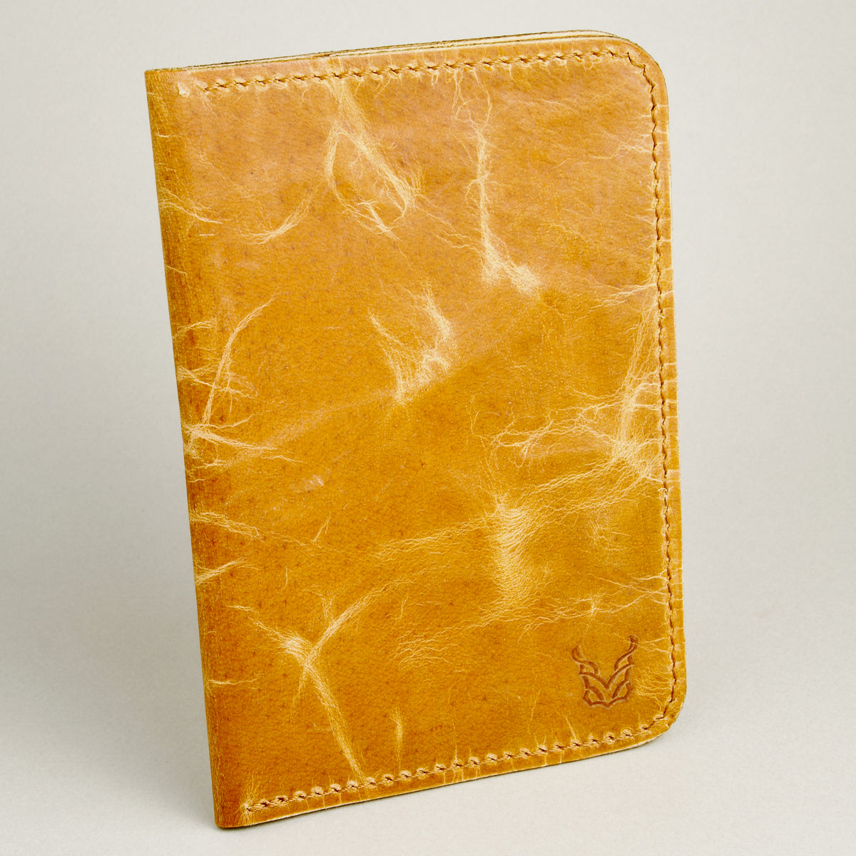 Tilted front view of vertical full-grain leather passport holder in amber, featuring handmade stitching and embossed logo. Markore Voyage Pass.